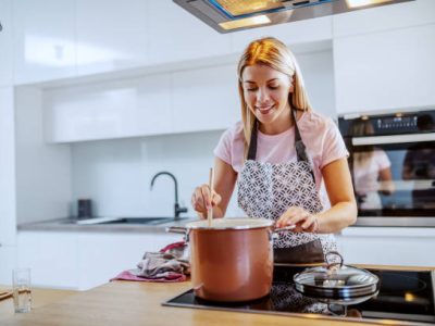 Beautiful smiling caucasian blond young woman in apron standing in kitchen and stirring soup in pot.